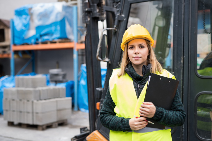 eine junge Frau in Arbeitsschutzkleidung steht vor einem Stapler im Außenbereich einer Produktionshalle